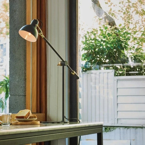 Adjustable arm black steel table lamp, lit beside a sunny window