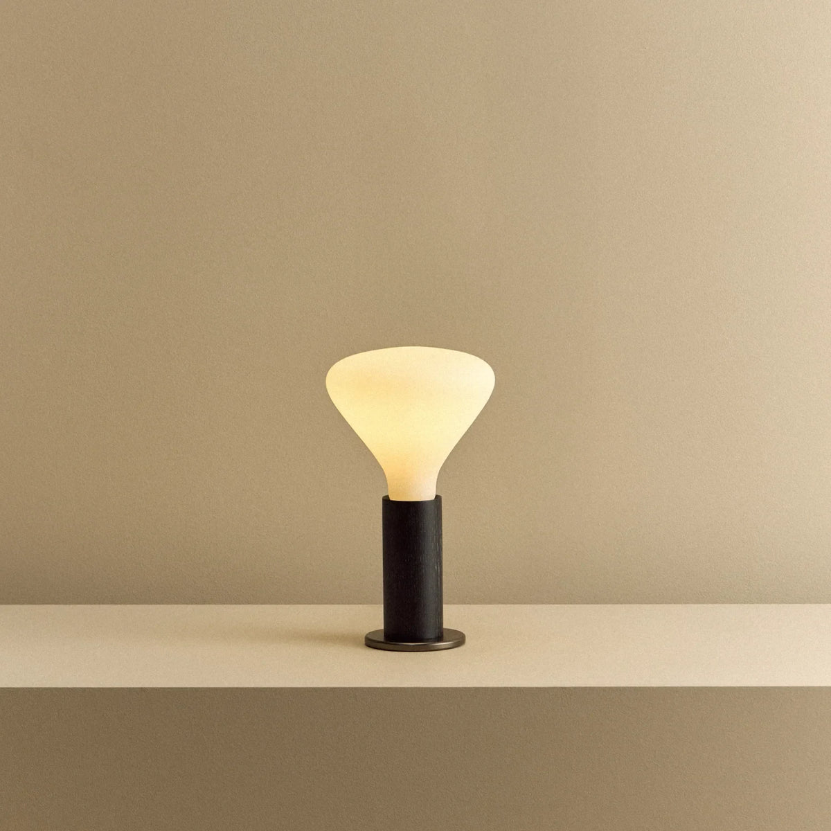Blackened oak table lamp with a flared cone opal bulb, lit, on a shelf
