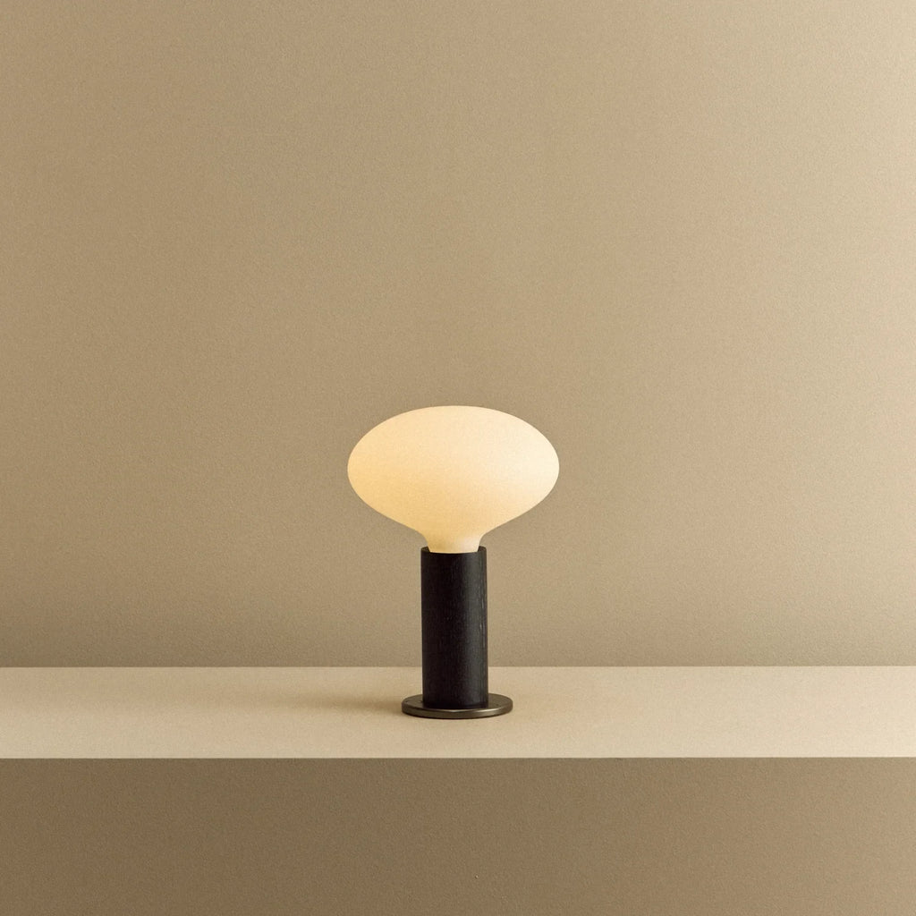 Blackened oak table lamp with a flat oval opal bulb, lit, on a shelf