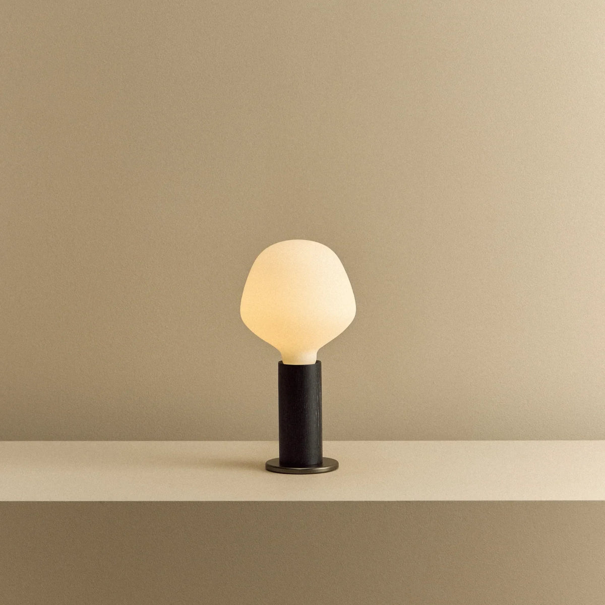 Blackened oak table lamp with a trapezoid opal bulb, lit, on a shelf