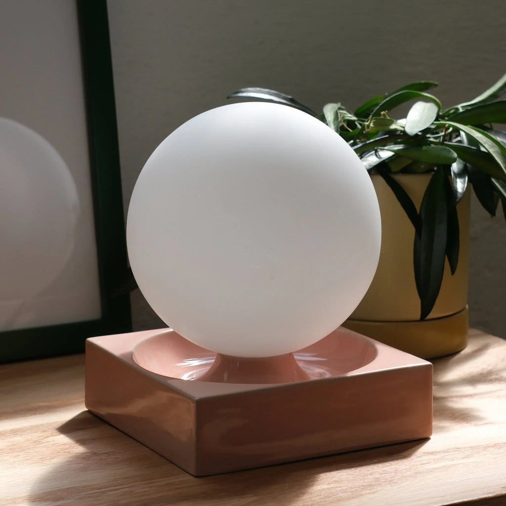 Ceramic cube table lamp with a blown glass globe, in pink beside a plant