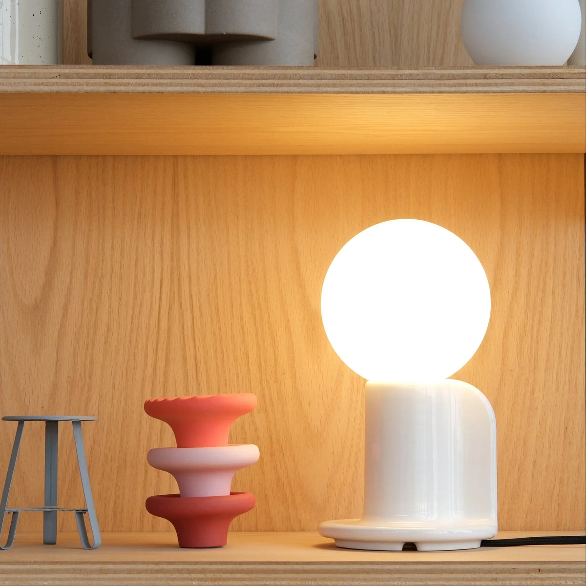 Ceramic table lamp with a paddle handle, in white, lit, on a shelf
