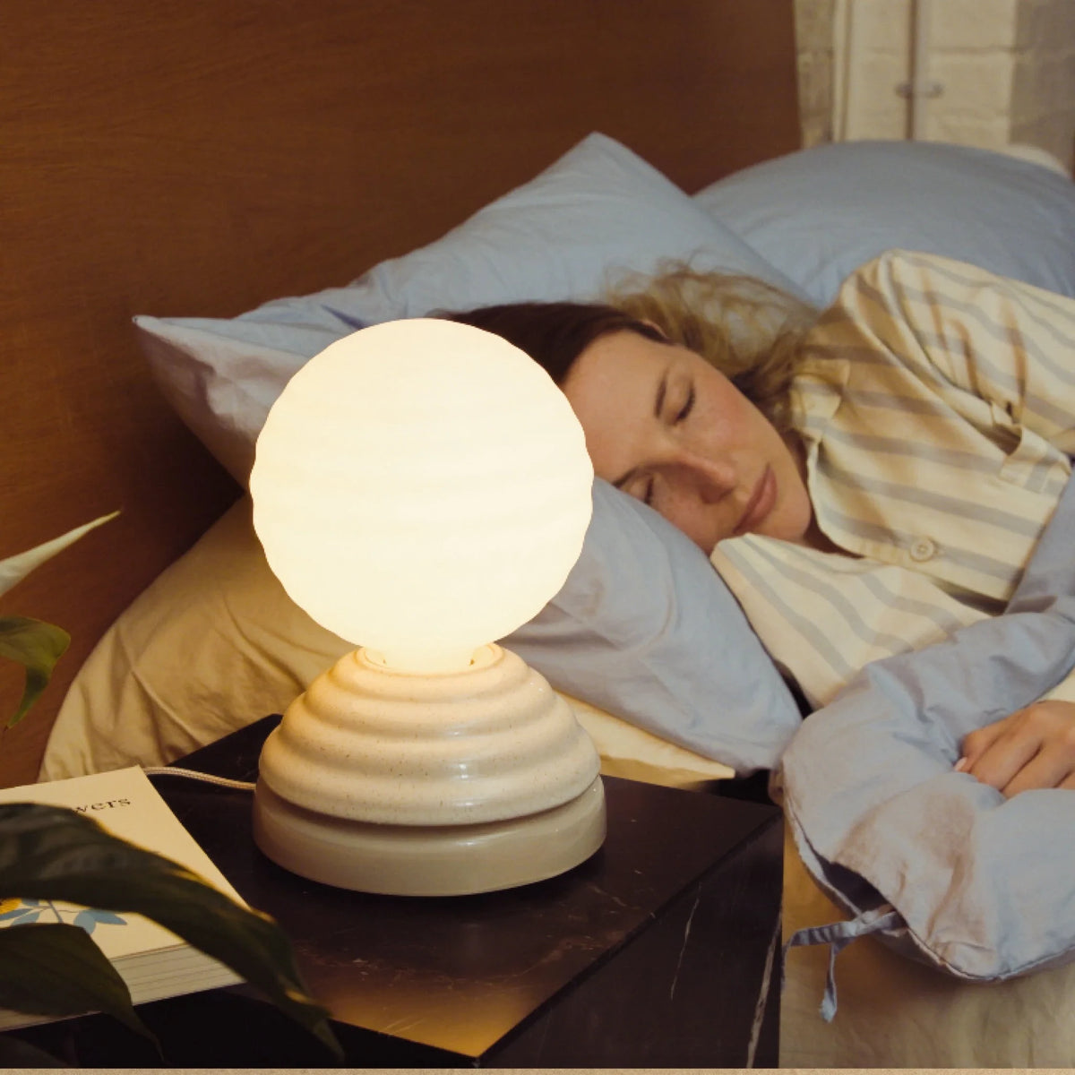 Ceramic wake light table lamp with sleep sounds, pebble white, beside a bed, lit