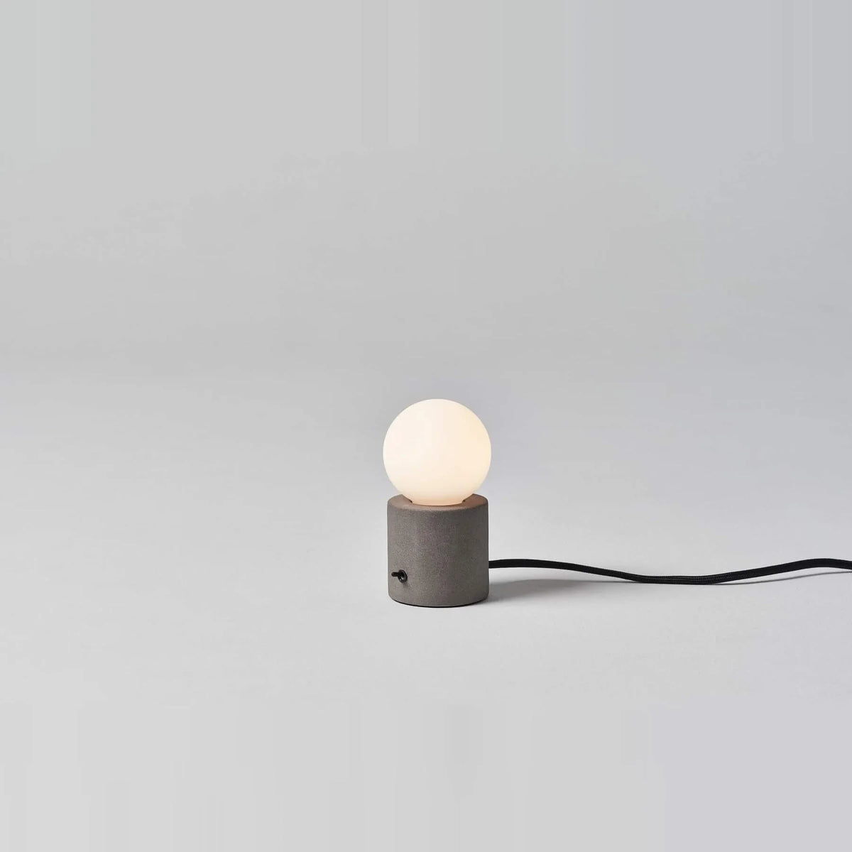 Concrete base table lamp with opal glass globe, lit, on a pale background with its cable