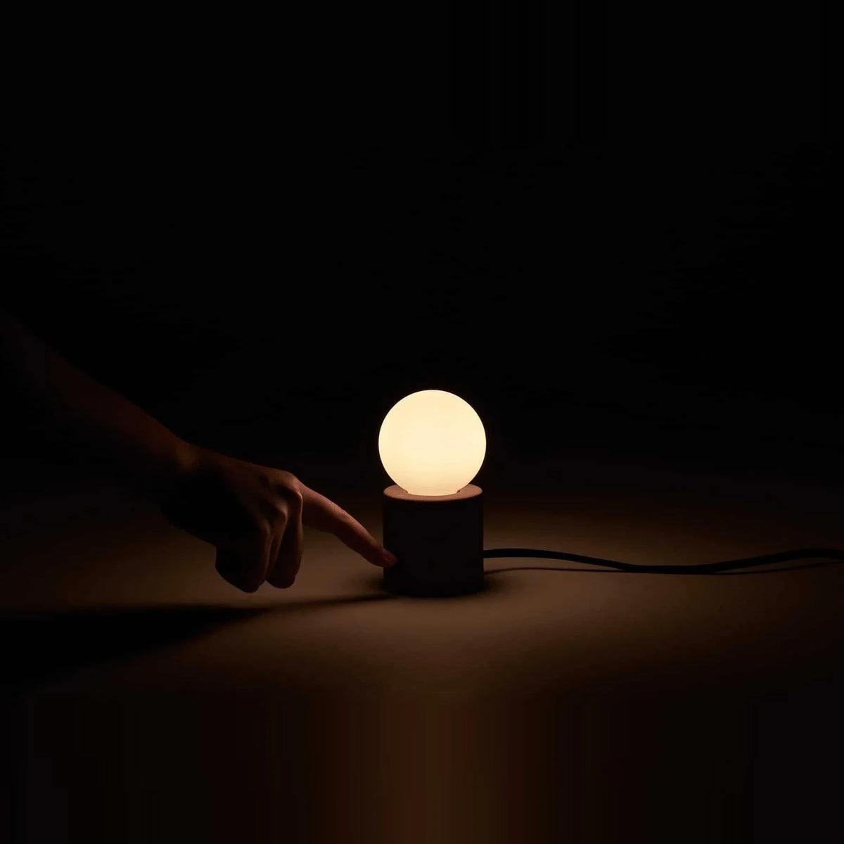 Concrete base table lamp with opal glass globe, lit in a dark room, a hand by the switch