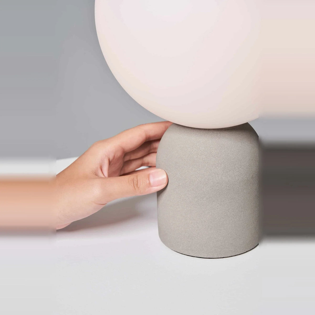 Concrete base table lamp with white glass globe, hand resting on the concrete base