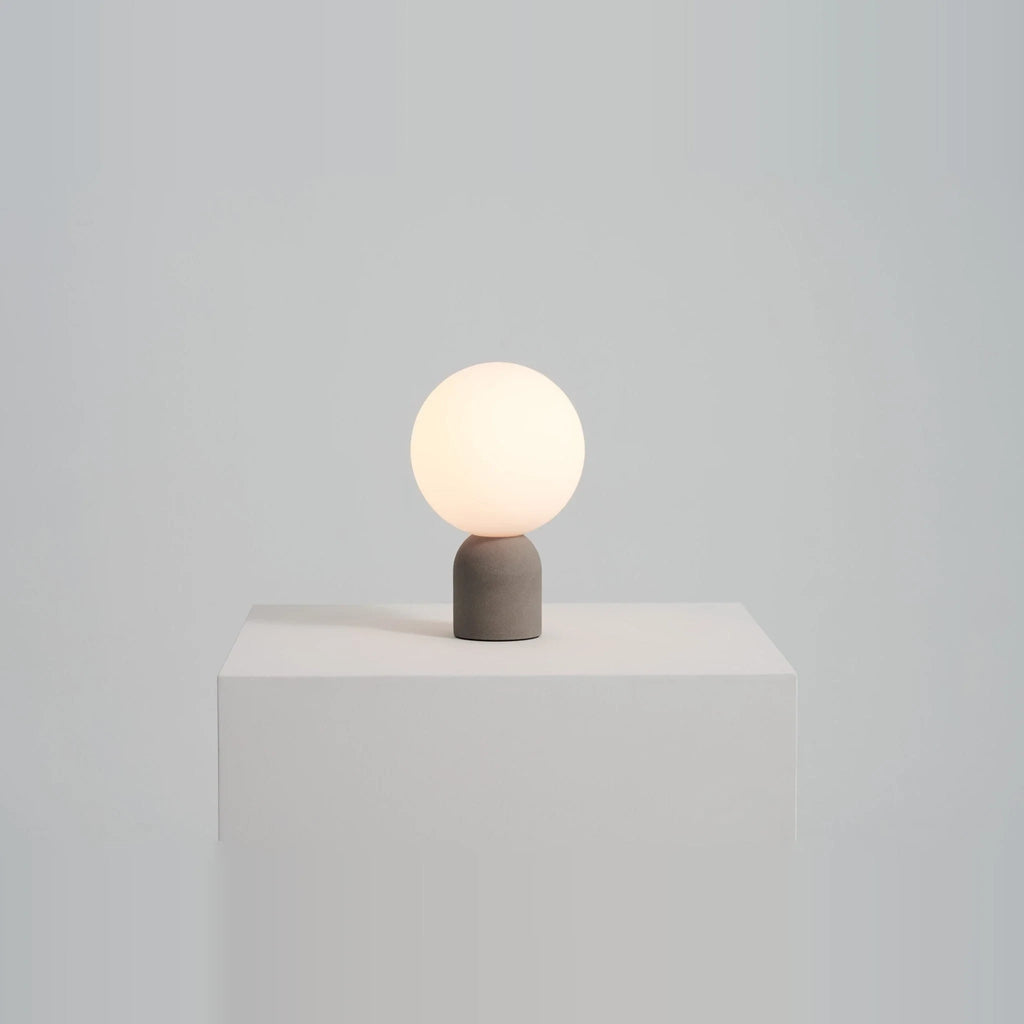 Concrete base table lamp with white glass globe, lit on a white plinth