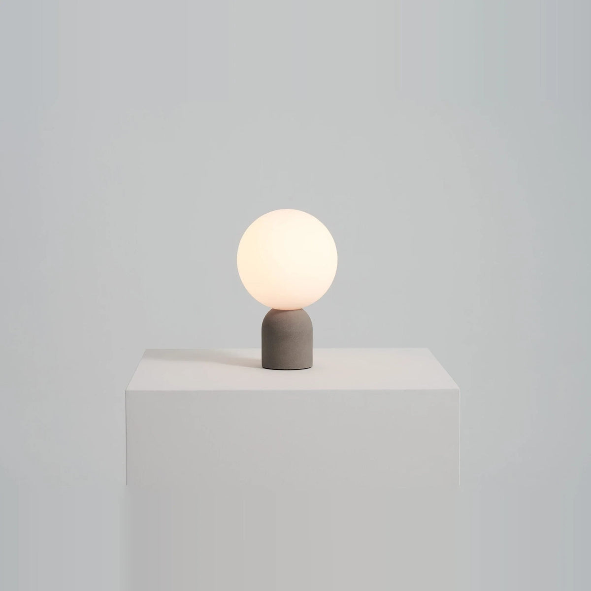 Concrete base table lamp with white glass globe, lit on a white plinth