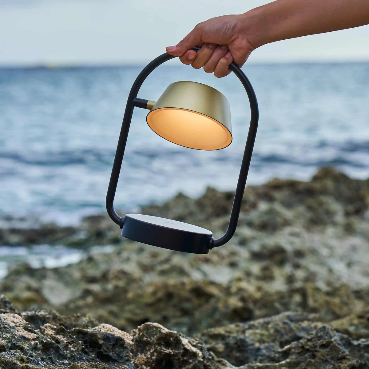 Cordless oval loop table lamp, champagne gold, carried by the handle on a rocky shore