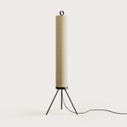 Cream linen tube floor lamp with black tripod base, against a plain wall