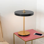 Disc-top LED table lamp with brass stem, in anthracite grey, with a phone charging on the table