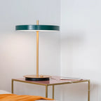 Disc-top LED table lamp with brass stem, in forest green, beside a bed