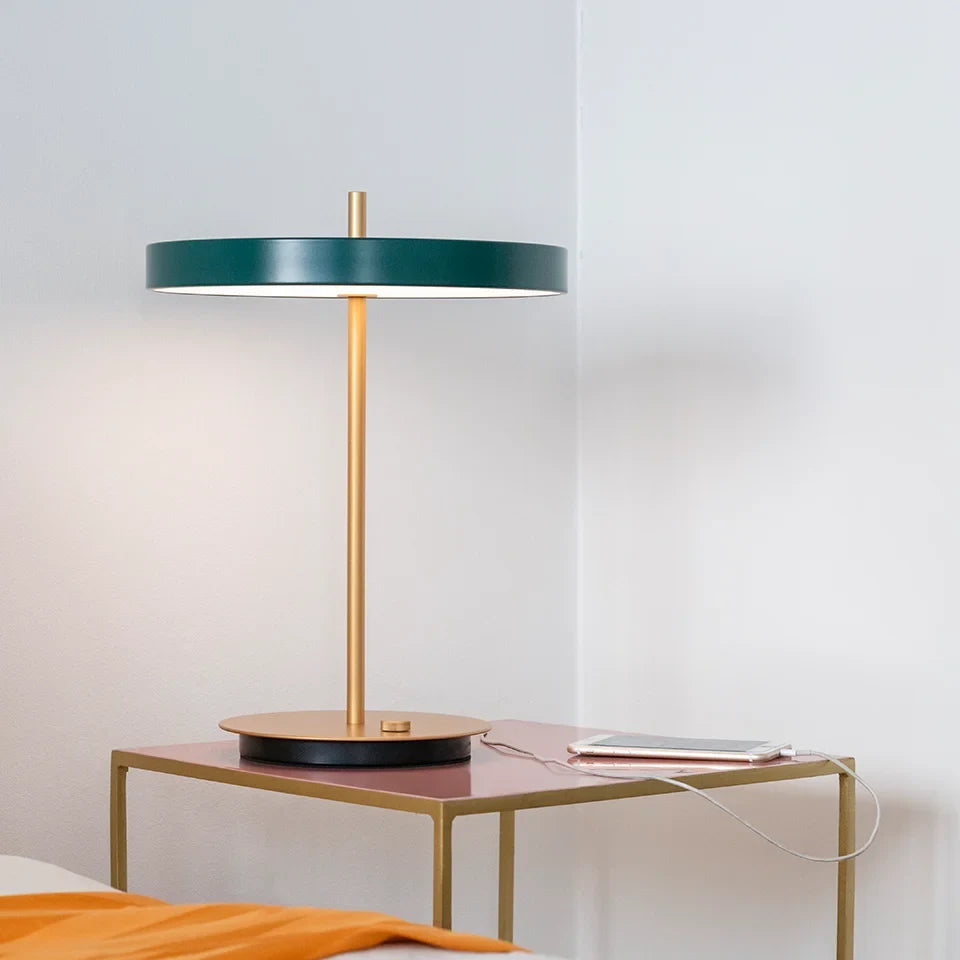 Disc-top LED table lamp with brass stem, in forest green, beside a bed