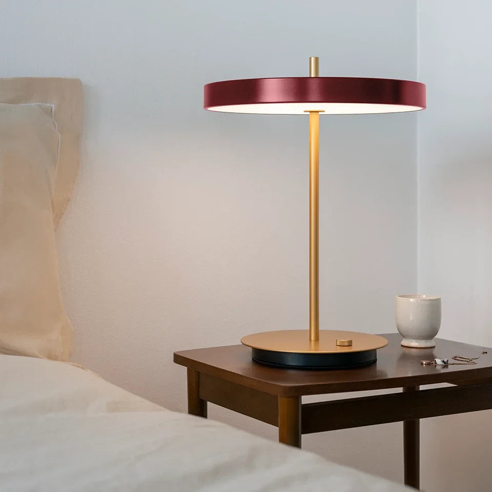 Disc-top LED table lamp with brass stem, in ruby red, beside a bed