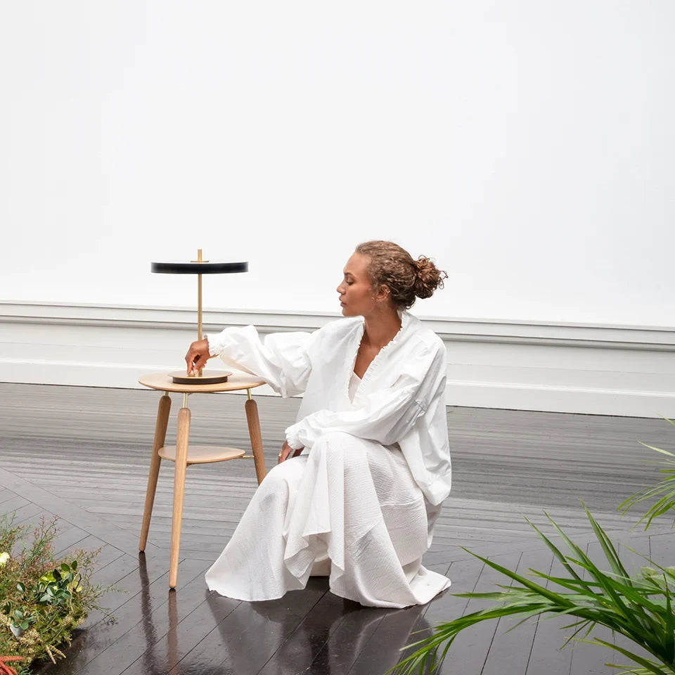Disc-top LED table lamp with brass stem, in black, on a stool beside a seated person