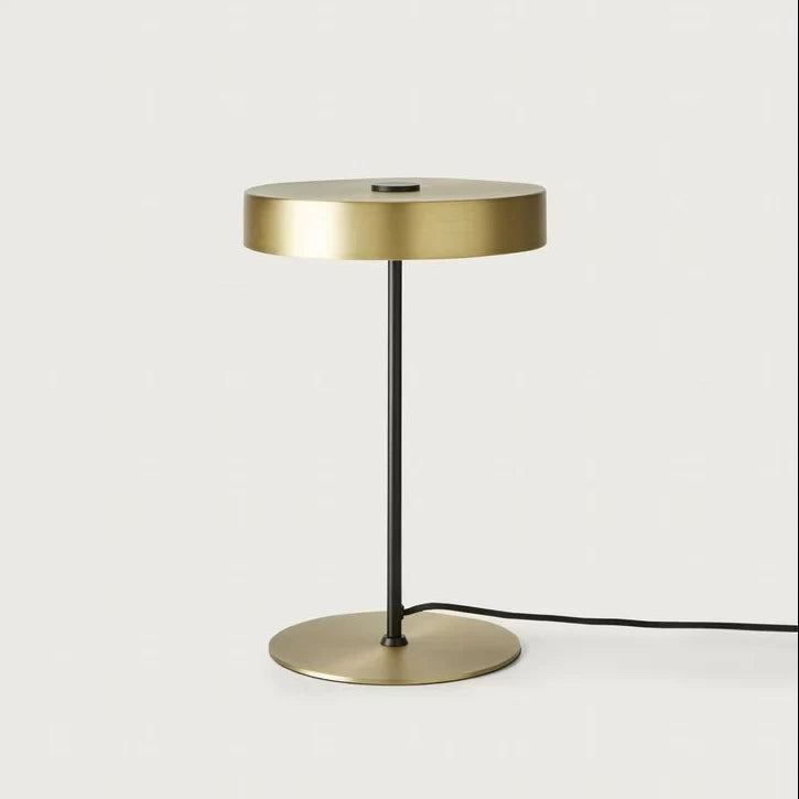 Flat disc shade LED table lamp on a slim stem, in matte brass, on a plain background