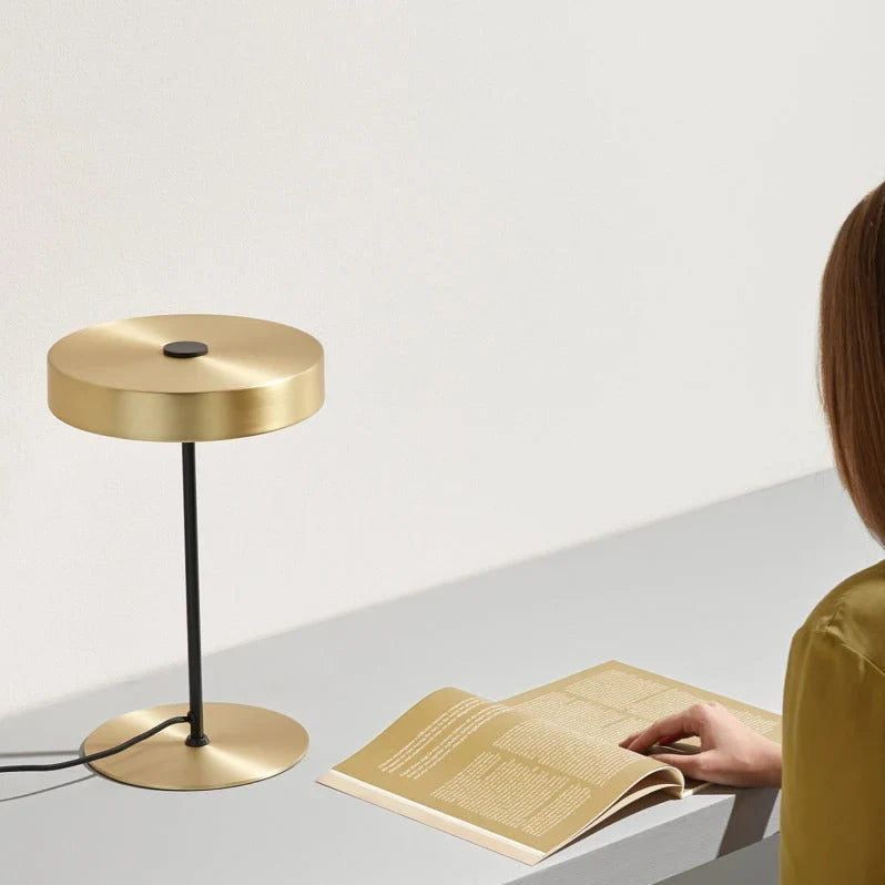 Flat disc shade LED table lamp on a slim stem, in matte brass on a desk beside an open book
