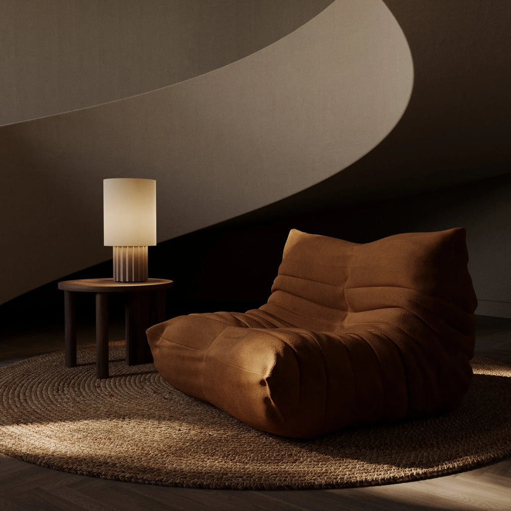 Fluted ceramic drum table lamp base, lit beside a lounge chair, with a shade (sold separately)