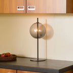 Mouth-blown glass globe table lamp on a slim steel stem, with smoke grey glass, lit, on a kitchen counter