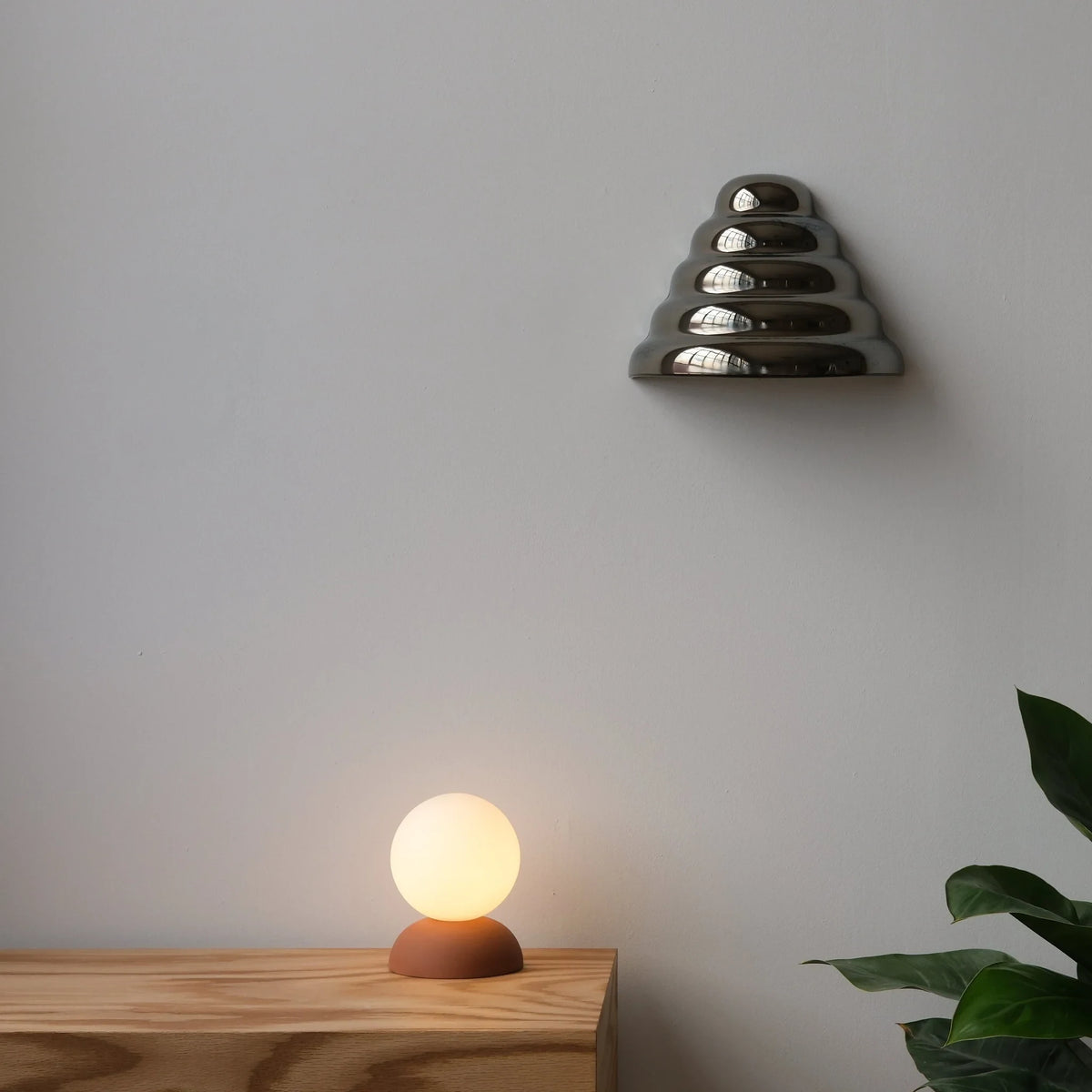 Glazed stoneware globe table lamp, in terracotta, lit, on a shelf beside a plant