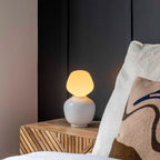 White porcelain table lamp with an angular opal bulb, on a slatted bedside cabinet beside a cushion