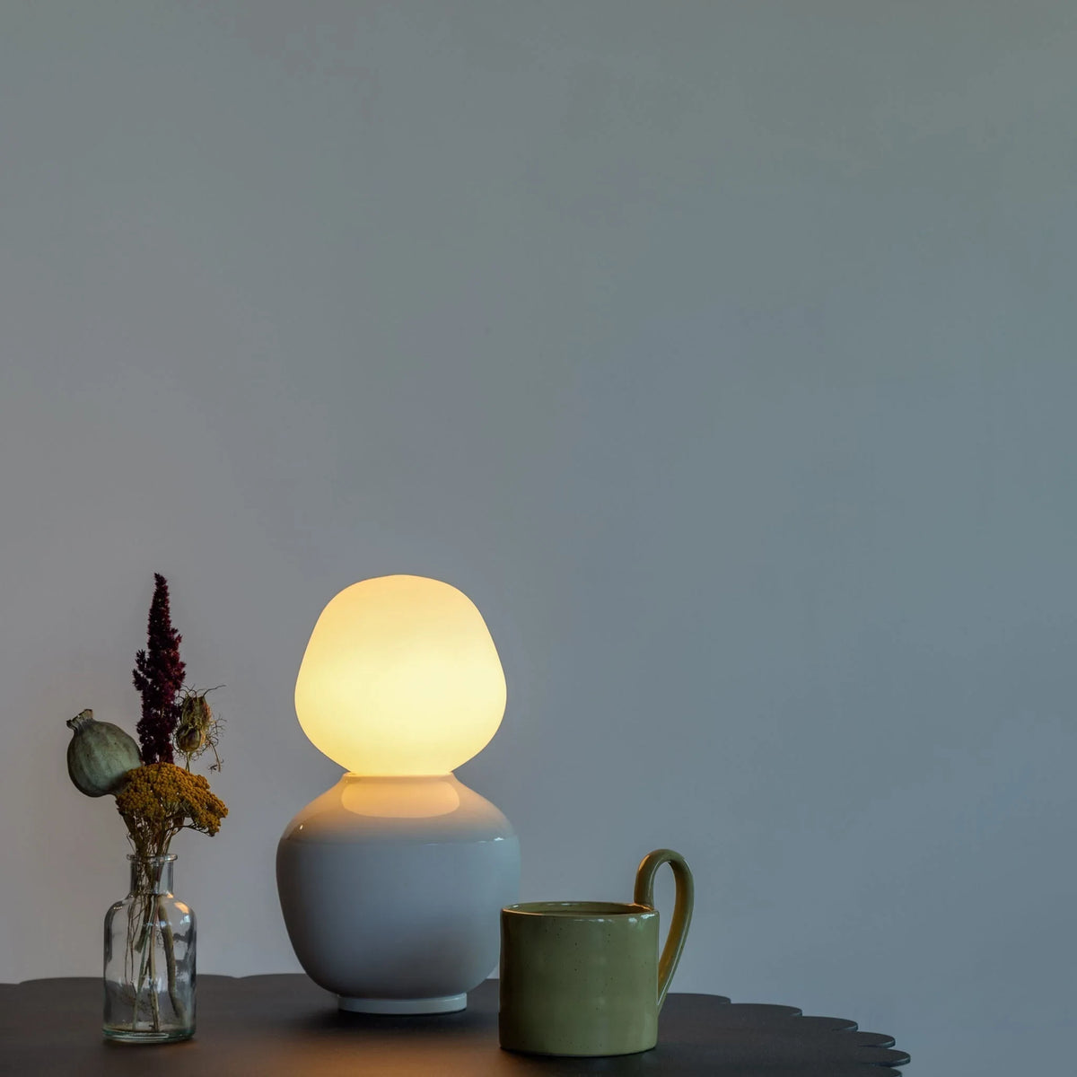 White porcelain table lamp with an angular opal bulb, beside dried flowers and a mug