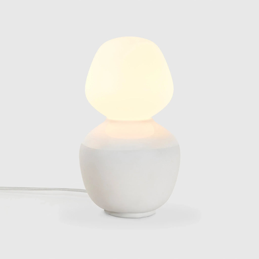 White porcelain table lamp with an angular opal bulb, lit, on a pale background
