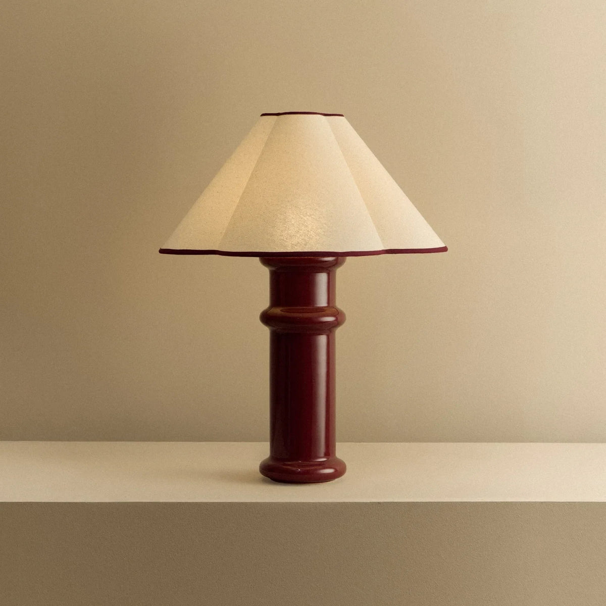 Ceramic table lamp, port red glaze with a folded cotton shade, lit, on a shelf