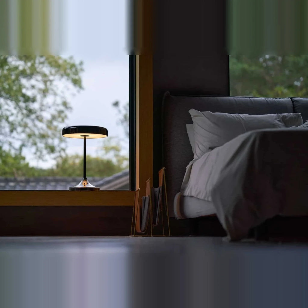 Black disc table lamp with rotating dimmer base, by a bed with a view out