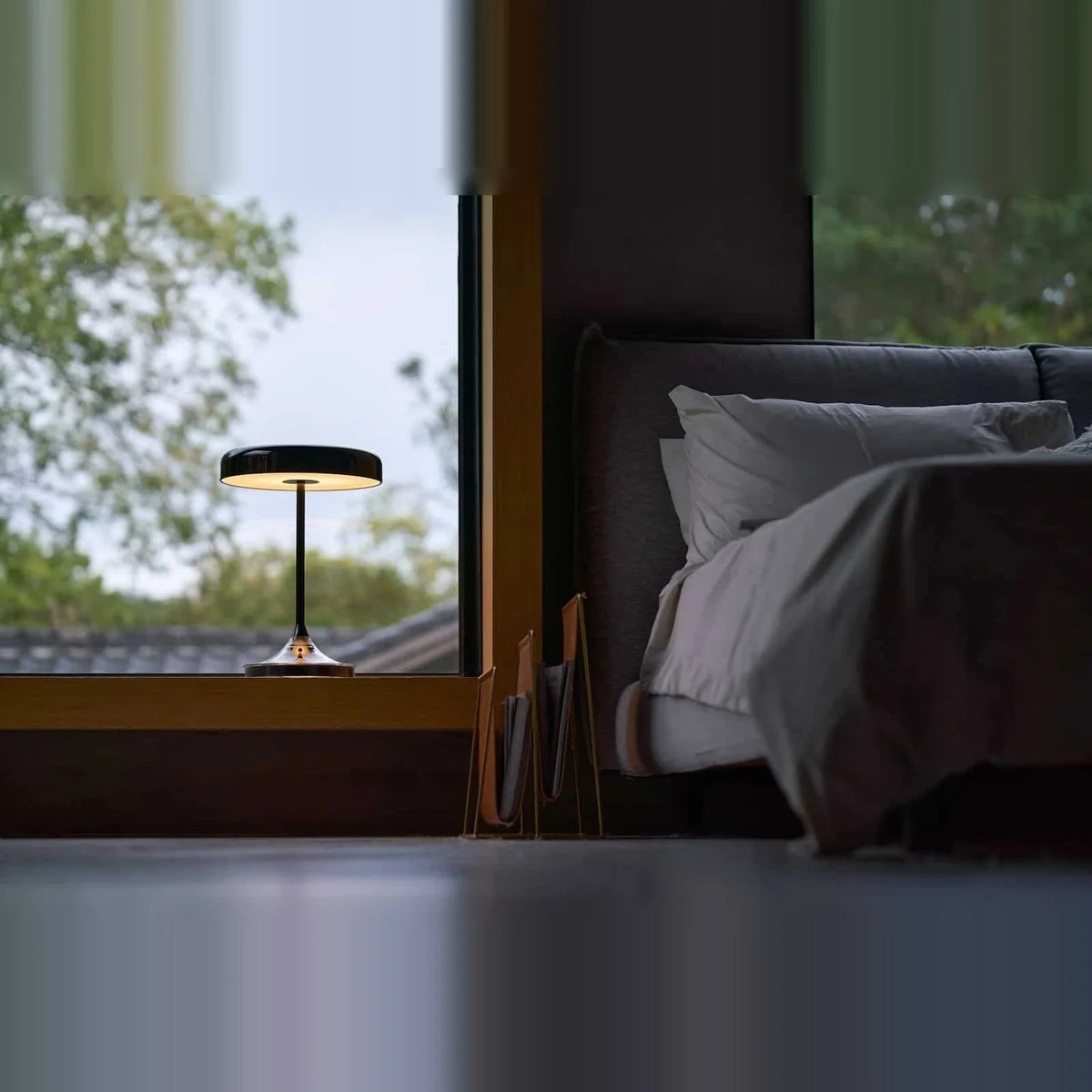 Black disc table lamp with rotating dimmer base, by a bed with a view out