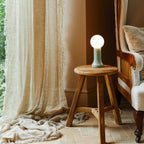 Mouth-blown glass table lamp in sea green, on a wooden stool beside a curtain