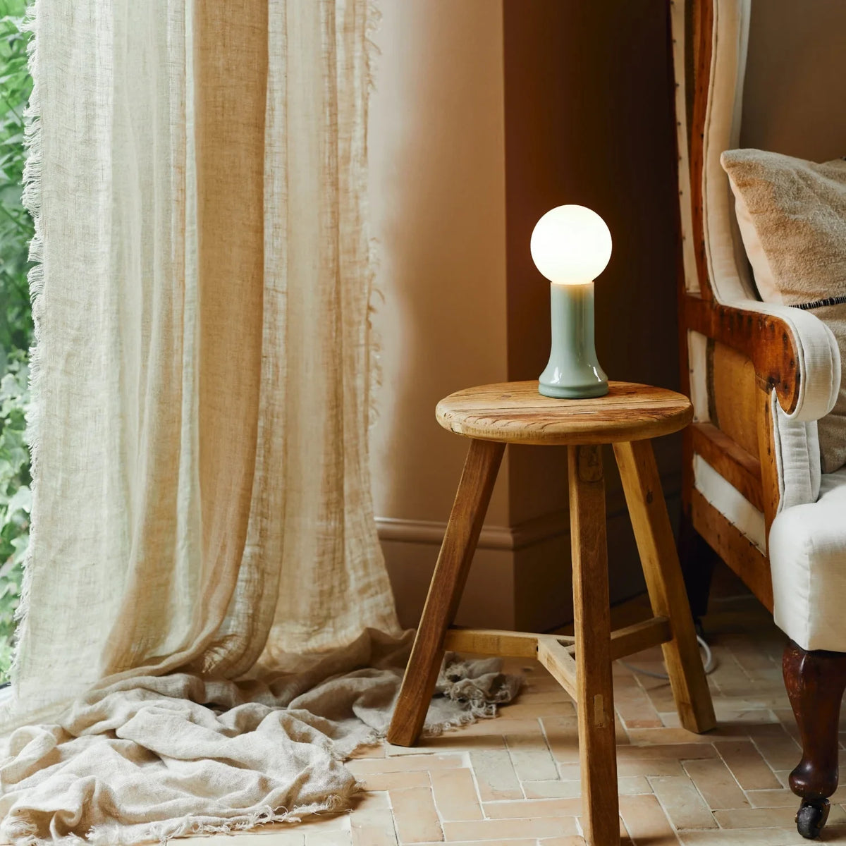 Mouth-blown glass table lamp in sea green, on a wooden stool beside a curtain
