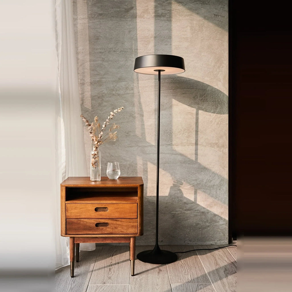 Slim disc LED floor lamp, in black beside a wooden cabinet