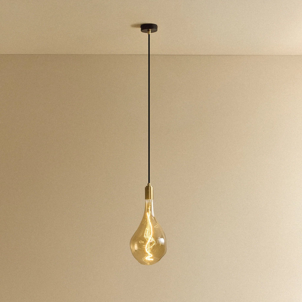 Tall tinted bulb pendant, in brass, lit, against a plain wall