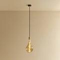 Tall tinted bulb pendant, in brass, lit, against a plain wall