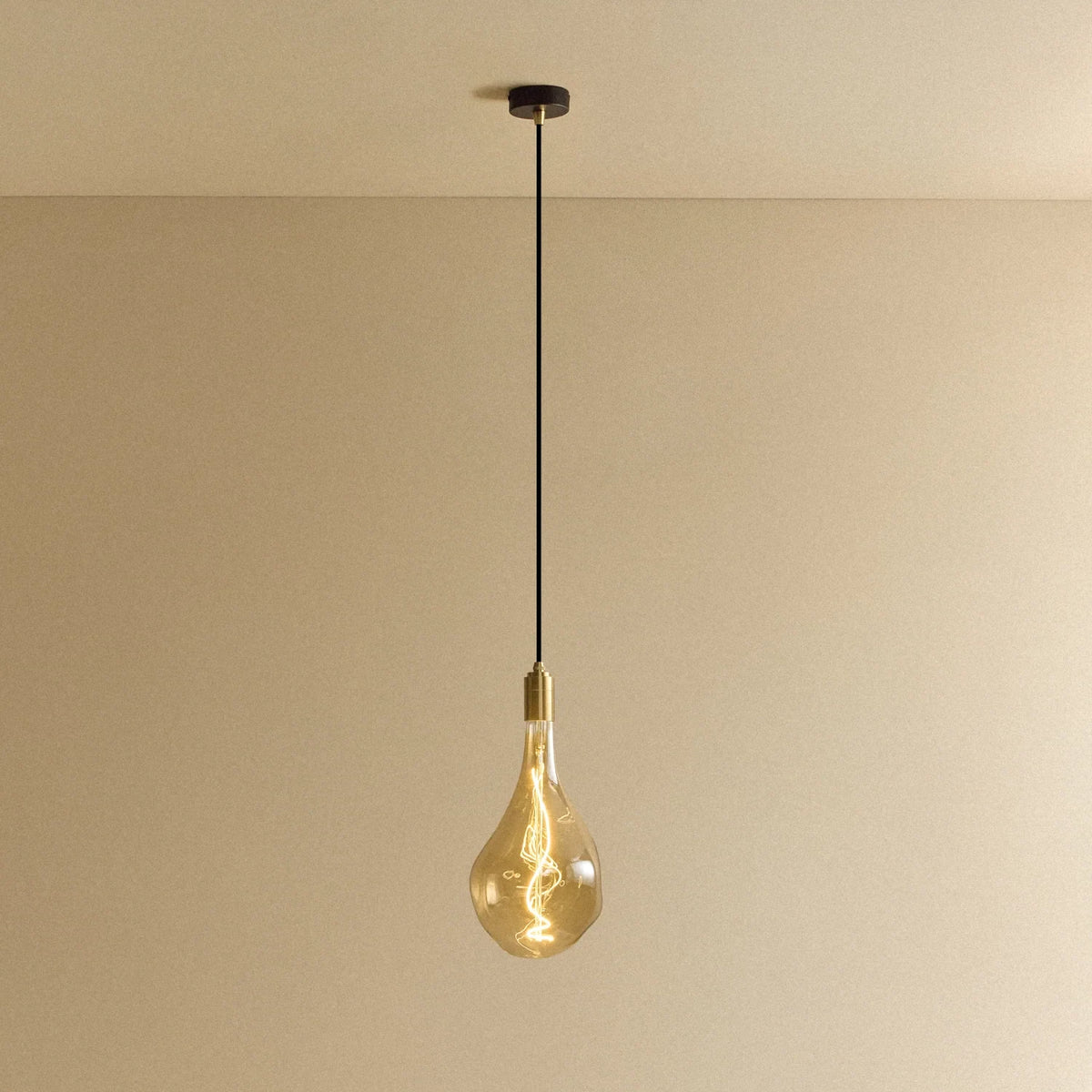 Tall tinted bulb pendant, in brass, lit, against a plain wall