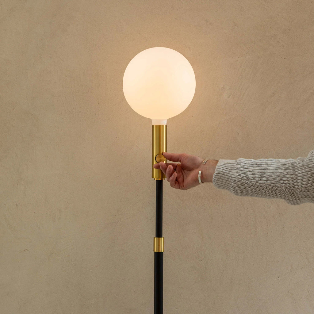 Brass telescopic floor lamp with a globe bulb, hand on the brass stem below the globe bulb