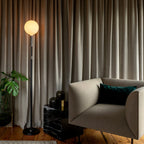 Brass telescopic floor lamp with a globe bulb, by curtains beside an armchair