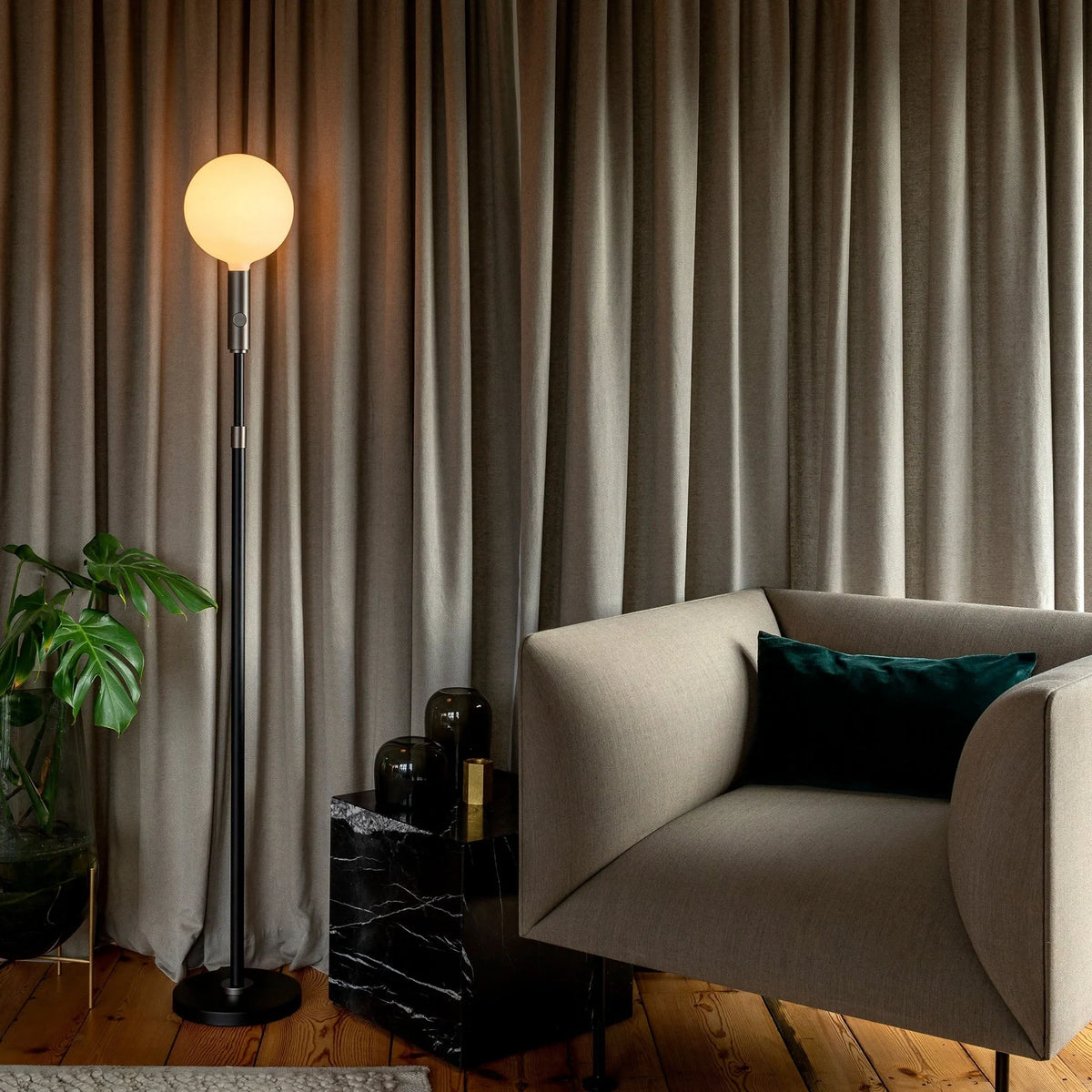 Brass telescopic floor lamp with a globe bulb, by curtains beside an armchair