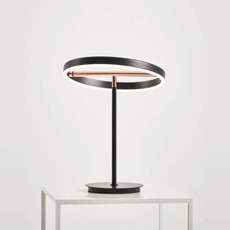 Tilting ring LED table lamp, black and copper, lit, on a white plinth