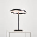 Tilting ring LED table lamp, black and copper, lit, on a white plinth