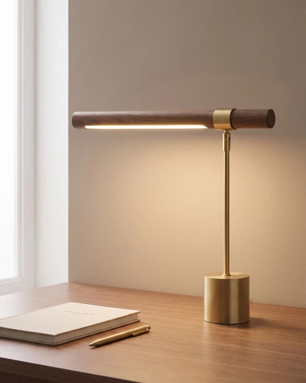 Walnut and brushed gold linear desk lamp, lit on a desk by a window