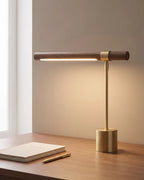 Walnut and brushed gold linear desk lamp, lit on a desk by a window