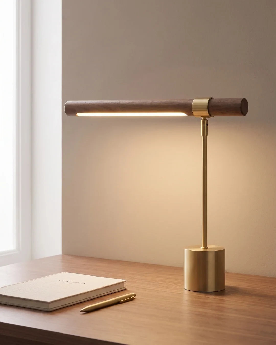 Walnut and brushed gold linear desk lamp, lit on a desk by a window