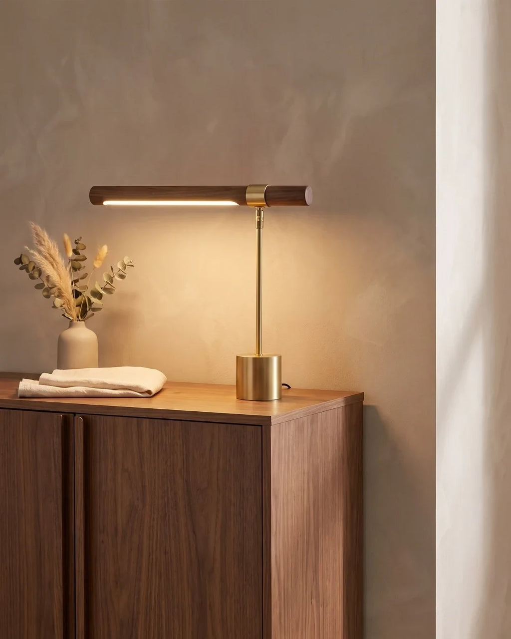 Walnut and brushed gold linear desk lamp, lit on a walnut cabinet