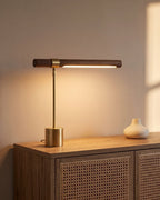 Walnut and brushed gold linear desk lamp, lit on a rattan sideboard