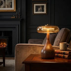Walnut-tone timber table lamp with a tinted glass disc, lit on a side table by a fireplace